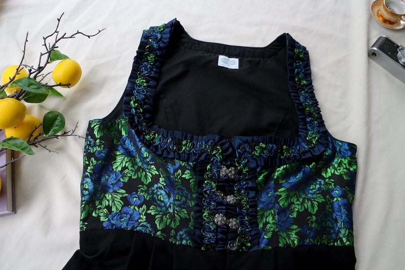 K&ouml;nnte beinhalten: Ein schwarzes Dirndl-Oberteil mit einem Blumenmuster in Blau und Gr&uuml;n. Das Oberteil hat einen quadratischen Ausschnitt und ist mit marineblauen R&uuml;schen verziert. Silberfarbene Verschl&uuml;sse zieren die Vorderseite.