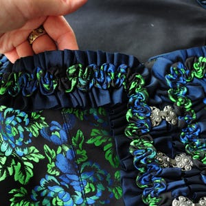 K&ouml;nnte beinhalten: Nahaufnahme eines dunkelblauen Kleidungsst&uuml;cks mit einem Blumenmuster in Blau und Gr&uuml;n. Das Kleidungsst&uuml;ck hat dekorative blaue und gr&uuml;ne R&uuml;schen und silberne Verschl&uuml;sse. Der Stoff hat einen Satin-Glanz.