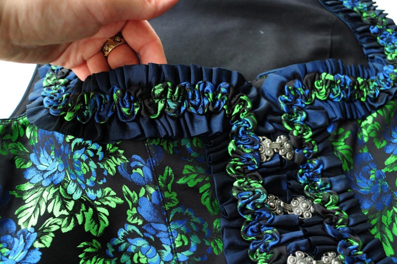 K&ouml;nnte beinhalten: Nahaufnahme eines dunkelblauen Kleidungsst&uuml;cks mit einem Blumenmuster in Blau und Gr&uuml;n. Das Kleidungsst&uuml;ck hat dekorative blaue und gr&uuml;ne R&uuml;schen und silberne Verschl&uuml;sse. Der Stoff hat einen Satin-Glanz.