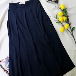 May include: A navy blue midi skirt featuring a high waistband with a label reading "JAPAN", vertical pleats, and a side closure with a row of dark buttons. The skirt is displayed alongside a vintage camera and a branch with yellow lemons.