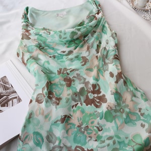 May include: A sleeveless dress with a cowl neckline, featuring a floral pattern in shades of green, brown, and beige. The dress is made of a lightweight, flowing fabric. A book with a black and white image is next to the dress.