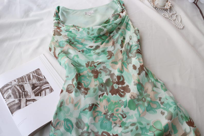 May include: A sleeveless dress with a cowl neckline, featuring a floral pattern in shades of green, brown, and beige. The dress is made of a lightweight, flowing fabric. A book with a black and white image is next to the dress.
