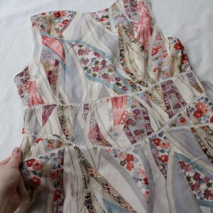 May include: A sleeveless dress with a patchwork design. The dress features a mix of floral and geometric patterns in shades of cream, pink, red, and blue. White trim accents the waist and neckline. A vintage camera is in the background.