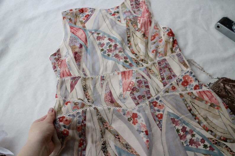 May include: A sleeveless dress with a patchwork design. The dress features a mix of floral and geometric patterns in shades of cream, pink, red, and blue. White trim accents the waist and neckline. A vintage camera is in the background.