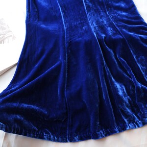 May include: A close-up of a vibrant blue velvet fabric, possibly a dress or skirt, with visible folds and a soft texture. The fabric is draped over a white surface, with a book visible in the background.