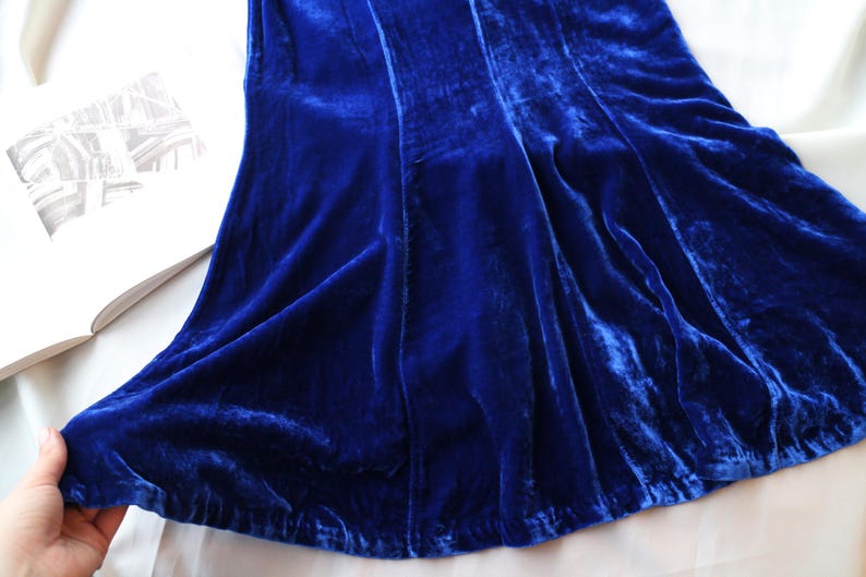 May include: A close-up of a vibrant blue velvet fabric, possibly a dress or skirt, with visible folds and a soft texture. The fabric is draped over a white surface, with a book visible in the background.