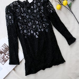 Puede incluir: Top negro de manga larga con intrincados adornos de lentejuelas y abalorios. El top presenta un cuello redondo y está adornado con lentejuelas plateadas en un patrón floral. La prenda se exhibe sobre una superficie blanca con limones y una cámara vintage.