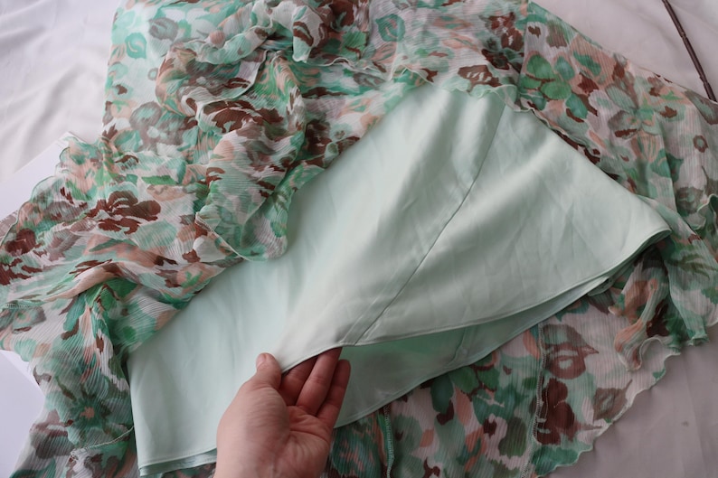 May include: A floral print dress with a light green lining. The outer layer features a pattern of green, brown, and peach-colored flowers on a sheer fabric. The dress is partially visible, with the lining and outer layer separated.
