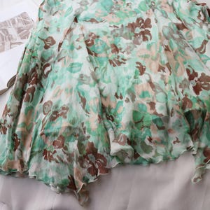 May include: A flowing, floral-print skirt in shades of green, brown, and cream. The fabric appears lightweight and semi-sheer, with a ruffled hemline. The skirt is draped over a book, suggesting a fashion or lifestyle theme.
