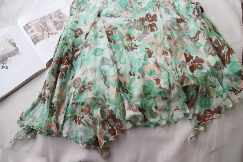 May include: A flowing, floral-print skirt in shades of green, brown, and cream. The fabric appears lightweight and semi-sheer, with a ruffled hemline. The skirt is draped over a book, suggesting a fashion or lifestyle theme.