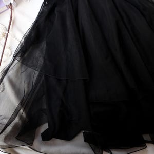 May include: A close-up of a black tulle skirt with multiple layers. The skirt is made of a sheer, lightweight fabric and is draped over a white surface. The skirt's layers create a flowing, voluminous effect.