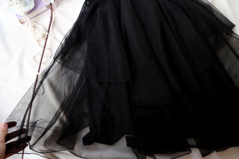 May include: A close-up of a black tulle skirt with multiple layers. The skirt is made of a sheer, lightweight fabric and is draped over a white surface. The skirt's layers create a flowing, voluminous effect.