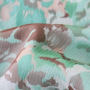 May include: Close-up of a fabric with a floral pattern. The design features a mix of colors, including light blue, brown, and peach, creating a soft, watercolor effect. The fabric appears to be lightweight and flowing.