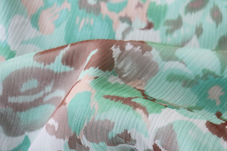 May include: Close-up of a fabric with a floral pattern. The design features a mix of colors, including light blue, brown, and peach, creating a soft, watercolor effect. The fabric appears to be lightweight and flowing.