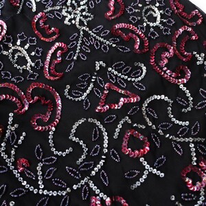 May include: Close-up of a black fabric adorned with intricate sequin and beadwork. The design features swirling paisley shapes and floral patterns in shades of red, silver, and white. The overall effect is elegant and detailed.