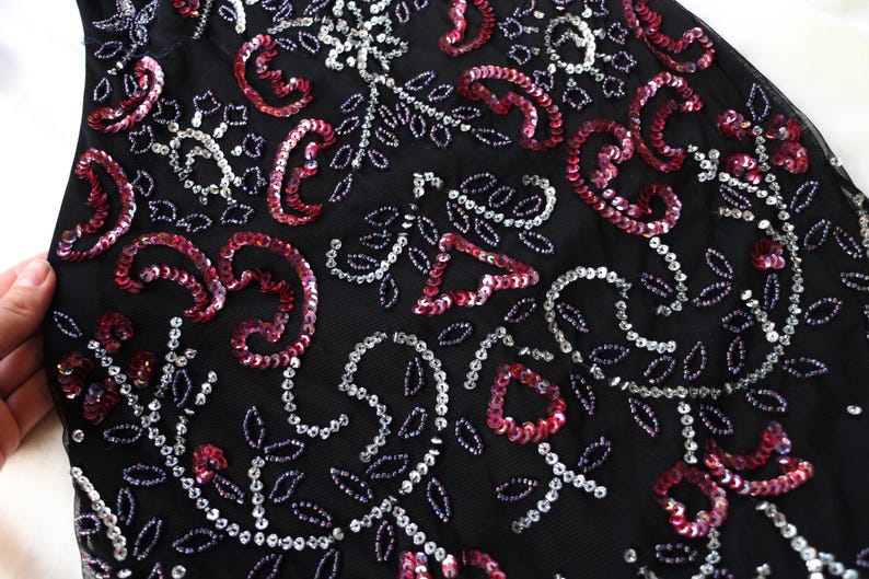May include: Close-up of a black fabric adorned with intricate sequin and beadwork. The design features swirling paisley shapes and floral patterns in shades of red, silver, and white. The overall effect is elegant and detailed.
