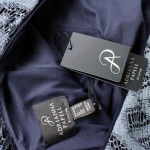 May include: Close-up of a navy blue evening dress with intricate lace detailing. The dress features a tag with the brand name "Adrianna Papell Evening". The dress is made of a dark blue fabric and has a zipper.