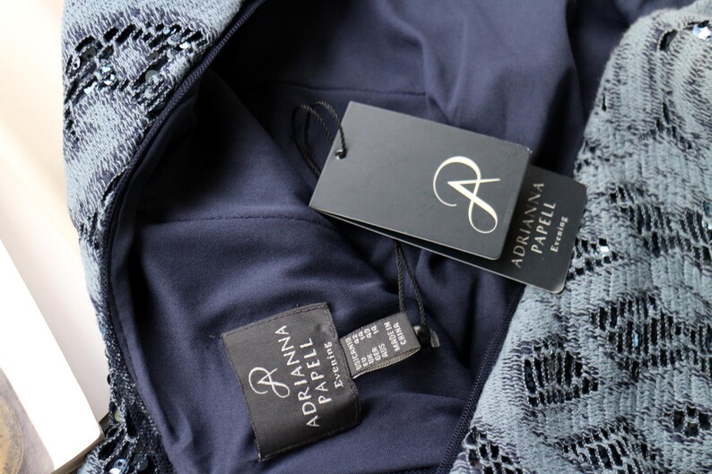 May include: Close-up of a navy blue evening dress with intricate lace detailing. The dress features a tag with the brand name "Adrianna Papell Evening". The dress is made of a dark blue fabric and has a zipper.