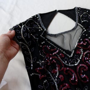 May include: Black sleeveless top with intricate sequin and bead embellishments. The top features a V-neckline and a keyhole back. Silver and red sequins create a floral pattern. The garment is held by a hand.