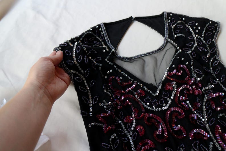 May include: Black sleeveless top with intricate sequin and bead embellishments. The top features a V-neckline and a keyhole back. Silver and red sequins create a floral pattern. The garment is held by a hand.