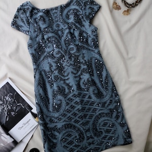 May include: A short-sleeved, blue sequined dress with an intricate, dark pattern. The dress is paired with a braided bracelet and gold-toned earrings. A magazine is visible in the background.