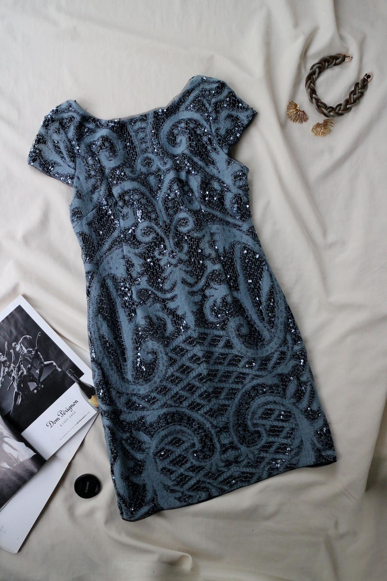 May include: A short-sleeved, blue sequined dress with an intricate, dark pattern. The dress is paired with a braided bracelet and gold-toned earrings. A magazine is visible in the background.