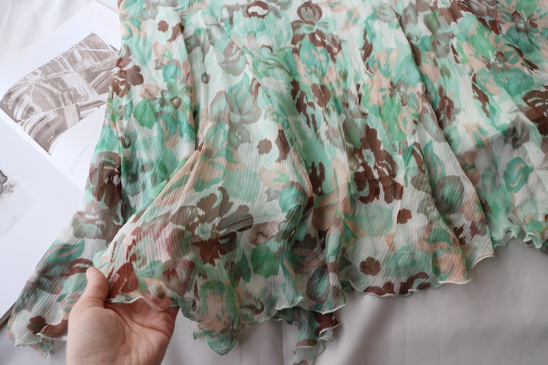 May include: A sheer, flowing garment with a floral pattern in shades of green, brown, and peach. The fabric appears lightweight and delicate, with a ruffled edge. The garment is likely a skirt or a dress.