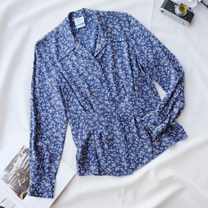 Op de afbeelding: Een blauwe blouse met bloemenprint met lange mouwen en een knoopsluiting aan de voorkant. De blouse heeft een kraag en een getailleerde taille. De stof is bedekt met een delicaat wit bloemenpatroon. De blouse ligt plat op een witte ondergrond.