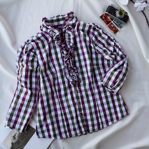 May include: A long-sleeved, button-down blouse in a gingham pattern of white, green, and purple. The blouse features a ruffled collar and cuffs, with a row of buttons down the front. The garment is laid flat on a white fabric surface.