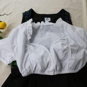 K&ouml;nnte beinhalten: Eine wei&szlig;e, kurz&auml;rmelige Bluse mit R&uuml;schenkanten und Knopfleiste. Die Bluse ist &uuml;ber einem schwarzen Kleid mit floralem Ausschnitt geschichtet. Gelbe Zitronen und eine Vintage-Kamera befinden sich im Hintergrund.