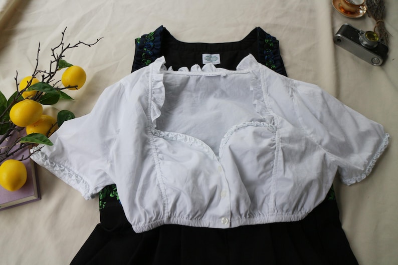 K&ouml;nnte beinhalten: Eine wei&szlig;e, kurz&auml;rmelige Bluse mit R&uuml;schenkanten und Knopfleiste. Die Bluse ist &uuml;ber einem schwarzen Kleid mit floralem Ausschnitt geschichtet. Gelbe Zitronen und eine Vintage-Kamera befinden sich im Hintergrund.