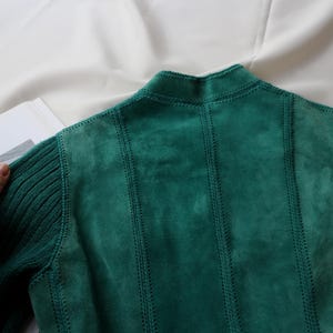 May include: A teal green suede jacket with ribbed knit sleeves and collar. The back of the jacket features vertical stitched panels. The jacket is displayed on a white surface.