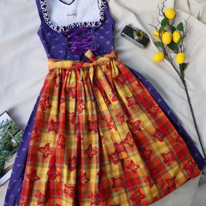 Könnte beinhalten: Ein traditionelles Dirndlkleid in Lila und Blumenmuster mit einem gelb-roten Karomuster. Das Oberteil hat eine Schnürung vorne und einen dekorativen Ausschnitt. Ein gelbes Band betont die Taille. Eine Vintage-Kamera und Zitronen sind in der Nähe.