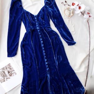 May include: A long, royal blue velvet dress with long sleeves and a button-down front. The dress features a V-neckline and a slight train. A white orchid and pearl necklace are placed near the dress on a white surface.