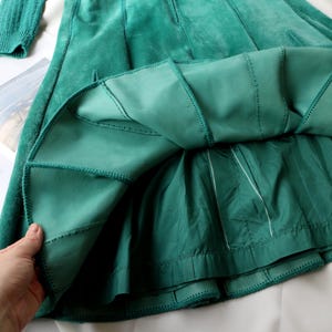 May include: A teal green suede skirt with a pleated design. The skirt has a fitted waist and a flared A-line shape. The lining is a lighter shade of green. The skirt is shown with a matching sleeve.