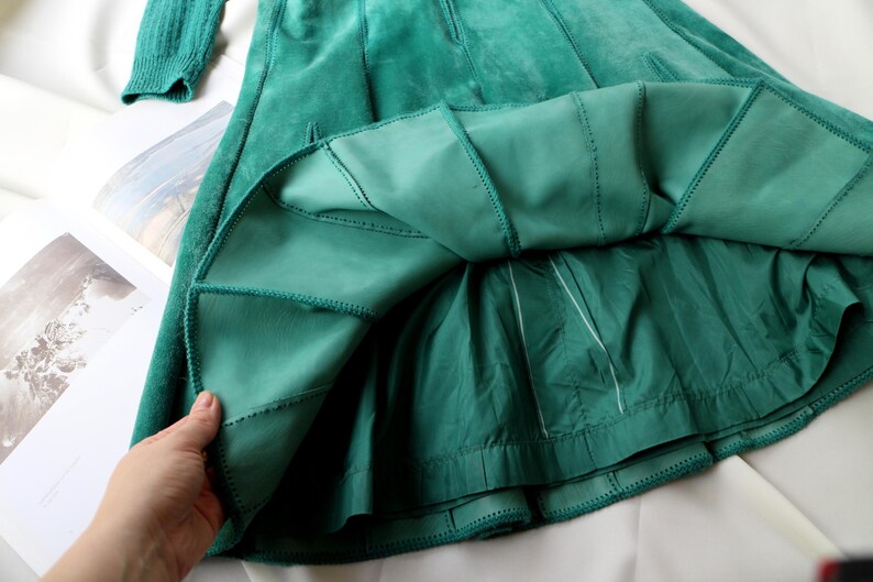 May include: A teal green suede skirt with a pleated design. The skirt has a fitted waist and a flared A-line shape. The lining is a lighter shade of green. The skirt is shown with a matching sleeve.