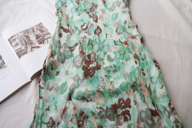 May include: A floral print dress in shades of green, brown, and white. The dress features a delicate pattern of flowers and leaves. The fabric appears lightweight and flowing, suggesting a summer garment. The dress is partially visible, with the top portion in focus.