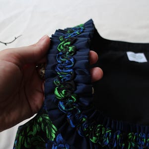 K&ouml;nnte beinhalten: Nahaufnahme eines marineblauen Kleidungsst&uuml;cks mit R&uuml;schenbesatz. Der Besatz hat gr&uuml;ne und blaue Akzente. Das Kleidungsst&uuml;ck hat ein Blumenmuster mit gr&uuml;nen und blauen Bl&auml;ttern. Der Hintergrund ist hell.