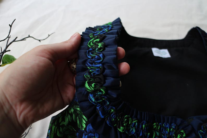 K&ouml;nnte beinhalten: Nahaufnahme eines marineblauen Kleidungsst&uuml;cks mit R&uuml;schenbesatz. Der Besatz hat gr&uuml;ne und blaue Akzente. Das Kleidungsst&uuml;ck hat ein Blumenmuster mit gr&uuml;nen und blauen Bl&auml;ttern. Der Hintergrund ist hell.