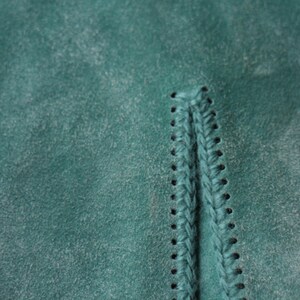 May include: Close-up of teal suede fabric with visible stitching and a braided detail. The texture of the material is slightly distressed, with a subtle, faded appearance. The stitching is a darker shade of teal.