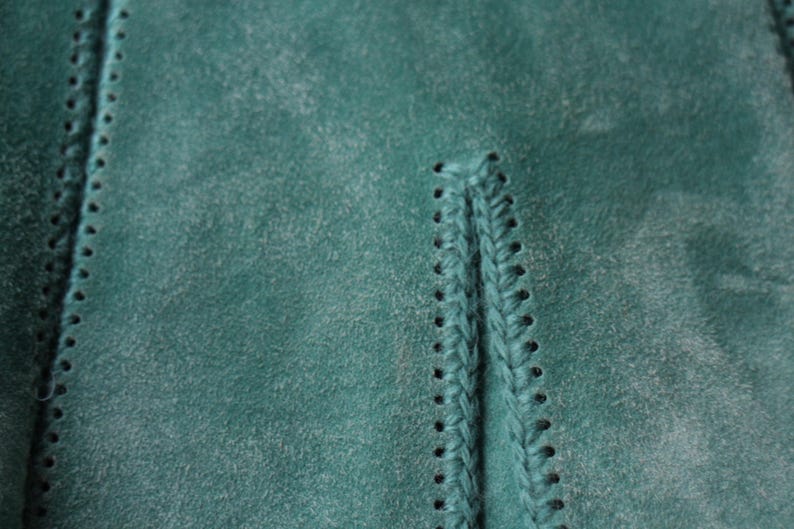 May include: Close-up of teal suede fabric with visible stitching and a braided detail. The texture of the material is slightly distressed, with a subtle, faded appearance. The stitching is a darker shade of teal.