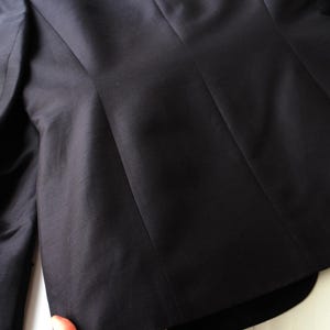 May include: Close-up of a black blazer, showcasing the tailored design and fabric texture. The image highlights the structured silhouette, with visible seams and a smooth, dark surface. The focus is on the back of the jacket, emphasizing its shape and construction.