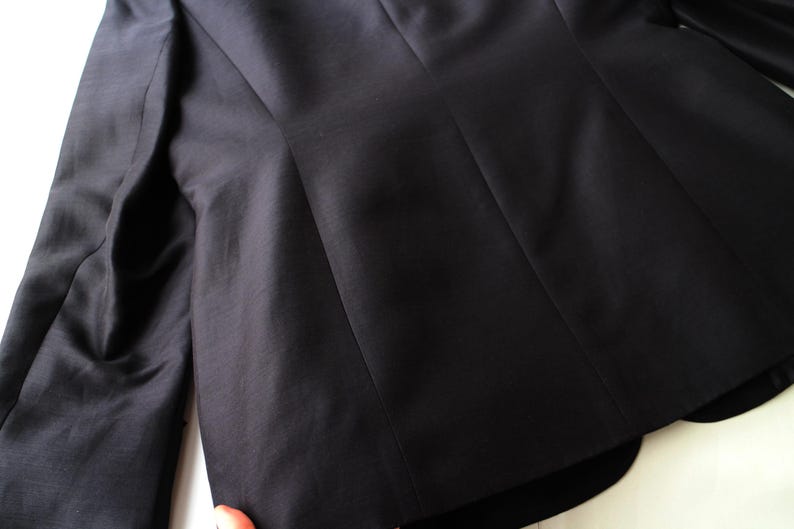 May include: Close-up of a black blazer, showcasing the tailored design and fabric texture. The image highlights the structured silhouette, with visible seams and a smooth, dark surface. The focus is on the back of the jacket, emphasizing its shape and construction.