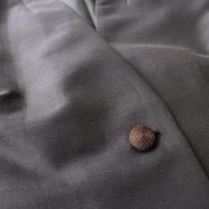 May include: Close-up of a dark gray blazer with a single, round, brown button. The fabric has a subtle sheen, and the button is made of tightly wound thread. The blazer's texture is smooth and appears to be a high-quality material.