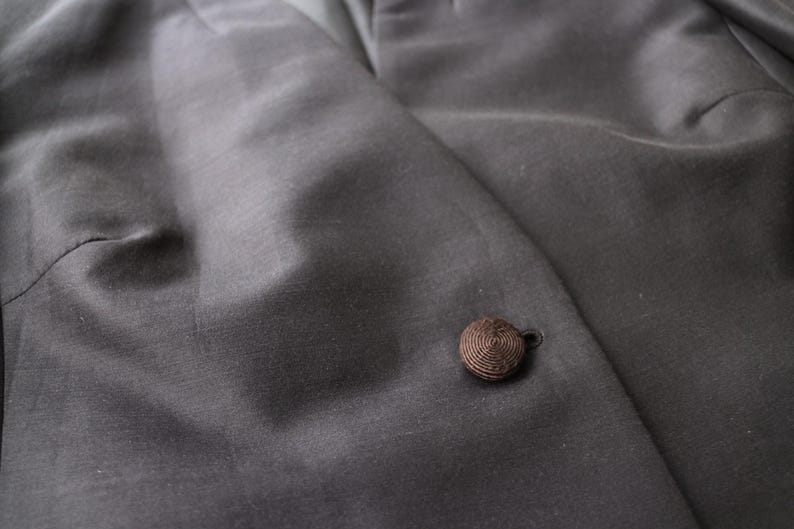 May include: Close-up of a dark gray blazer with a single, round, brown button. The fabric has a subtle sheen, and the button is made of tightly wound thread. The blazer's texture is smooth and appears to be a high-quality material.