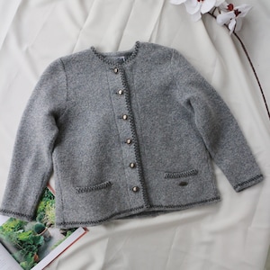 May include: A grey wool jacket with long sleeves, a round neckline, and a button-up front. The jacket has two front pockets and is trimmed with dark grey stitching. The buttons are gold-coloured. A fashion item.
