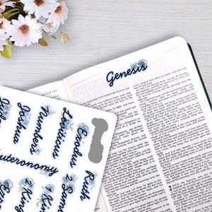 May include: A sheet of adhesive Bible index tabs featuring blue floral accents and black cursive text for book names like Genesis, Exodus, and Ruth. The tabs are displayed alongside an open Bible with one tab already applied to the page.
