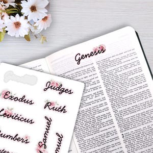 May include: A sheet of white adhesive stickers featuring black cursive text for Bible book names like Genesis, Exodus, and Judges, each accented with small pink floral illustrations. The stickers are displayed next to an open Bible page.