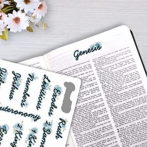 May include: A sheet of adhesive Bible index tabs featuring black cursive text on a blue floral background, including labels for Genesis, Exodus, Leviticus, Numbers, Deuteronomy, Joshua, 1 Samuel, 2 Samuel, and 1 Kings, placed on an open Bible.