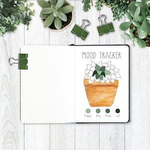 May include: Open notebook with a watercolor illustration labeled "MOOD TRACKER." The drawing features a succulent in a terracotta pot, with a color-coded mood scale below. Green binder clips and potted succulents surround the notebook on a white wood surface.